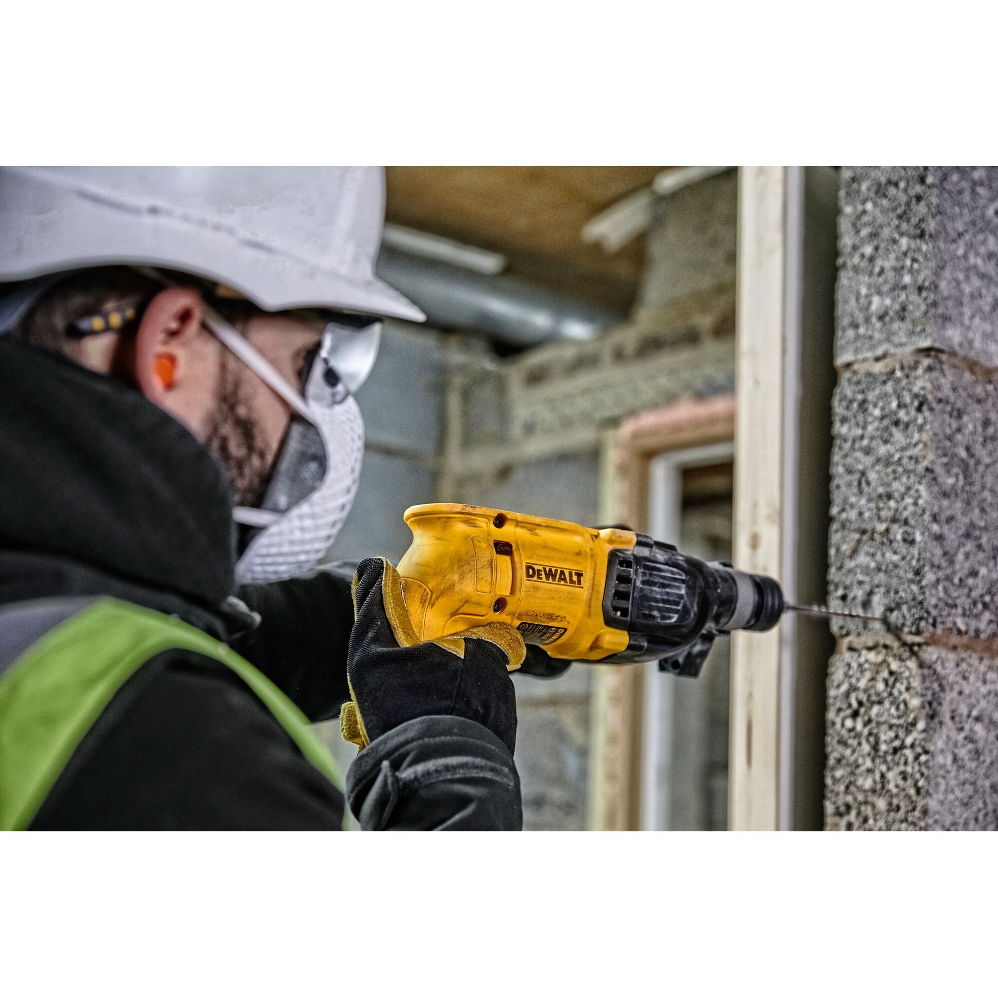 A person wearing protective gear uses a yellow DEWALT hammer drill (model D25033K) to drill into a concrete block wall on a construction site.