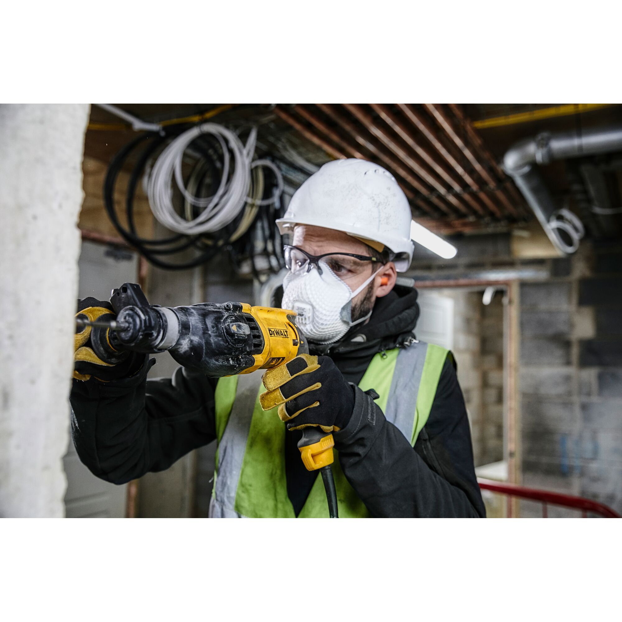 A person wearing a white safety helmet, gloves, and a high-visibility vest is using a DEWALT D25133K rotary hammer drill on a concrete wall at a construction site.