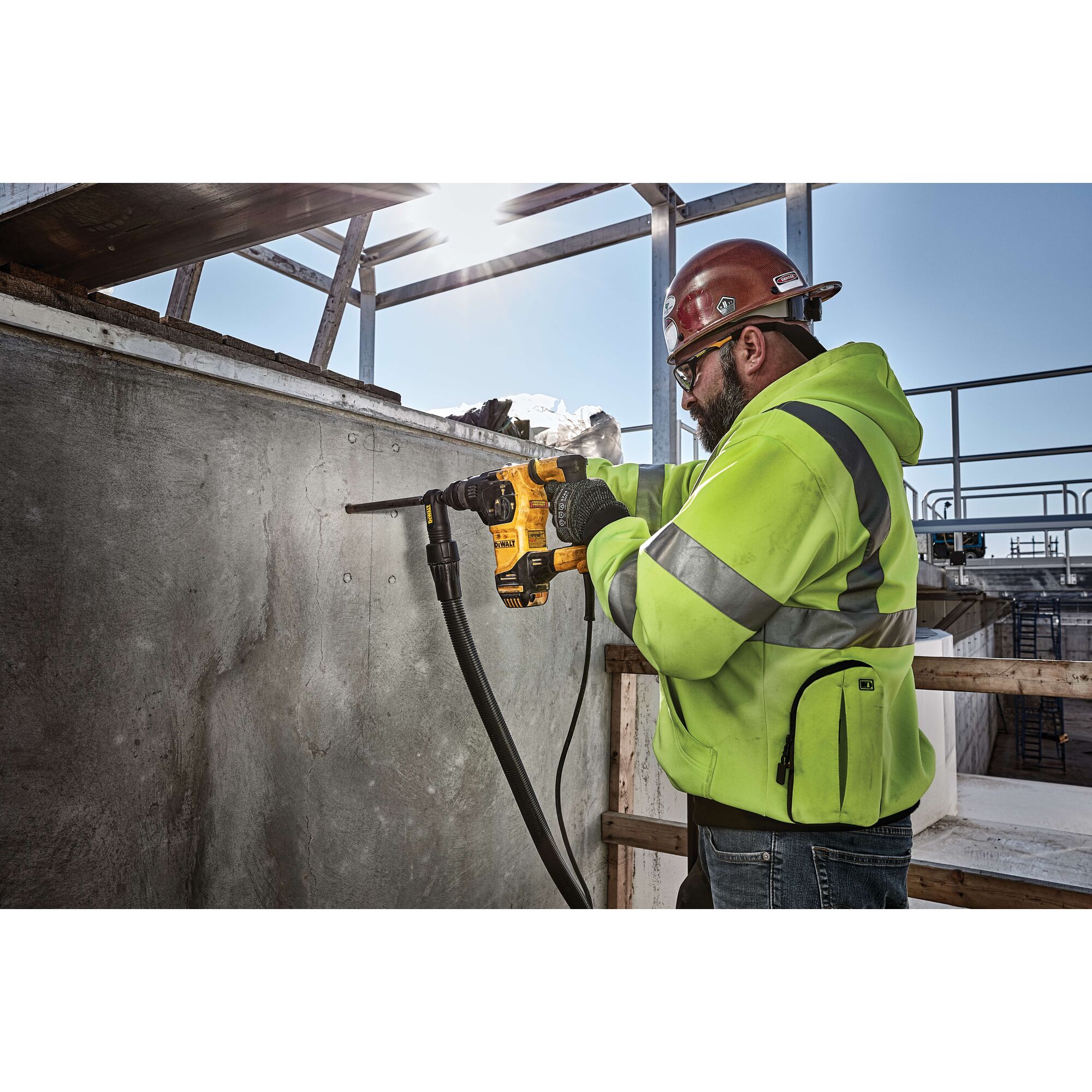 Person wearing a high-visibility jacket and hard hat using a DEWALT rotary hammer drill to drill into a concrete wall at a construction site.