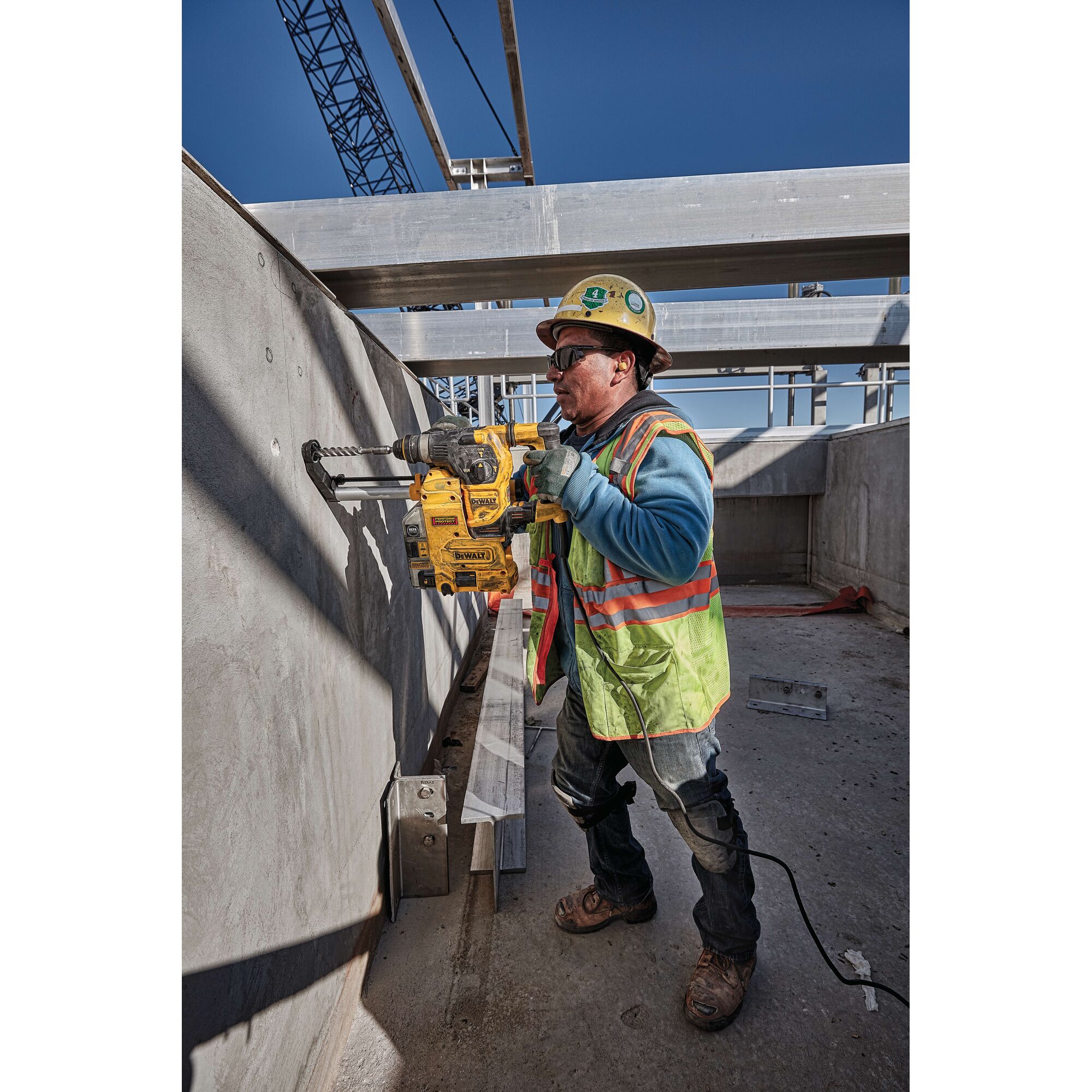 A construction worker wearing a safety vest, helmet, and gloves is operating a DEWALT rotary hammer drill (model D25333K) to drill into a concrete wall on an outdoor construction site, surrounded by metal beams and structural elements. The worker’s face is intentionally blurred.