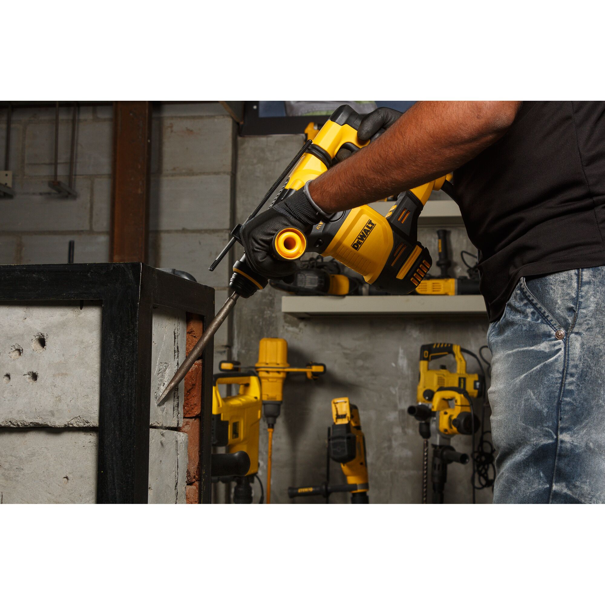 A person wearing gloves uses a DEWALT rotary hammer drill to chisel concrete blocks in a workshop. Other yellow DEWALT power tools are visible in the background.