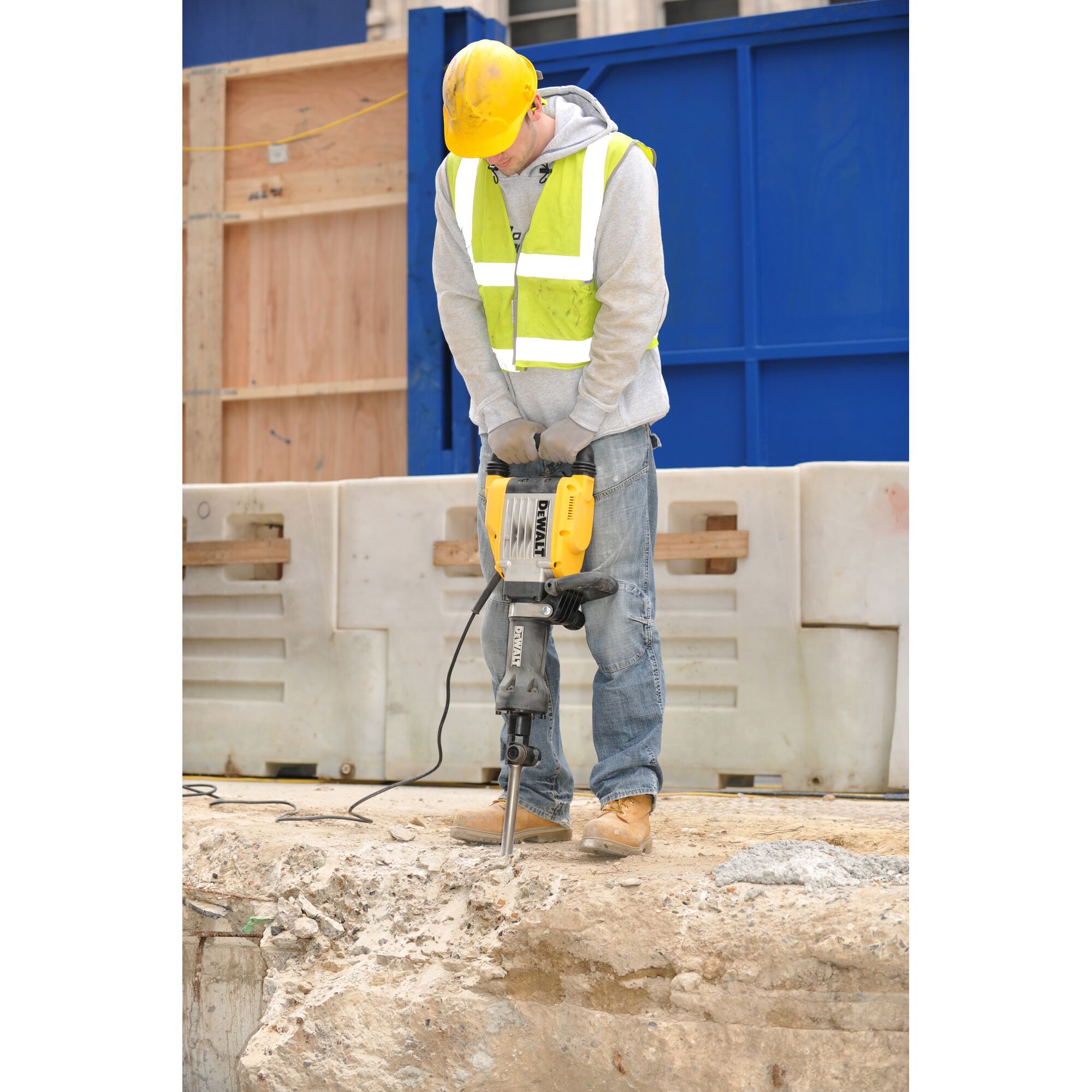 A construction worker wearing a yellow hard hat, reflective safety vest, and gloves is using a DEWALT electric jackhammer (model D25961K) to break concrete at a construction site.