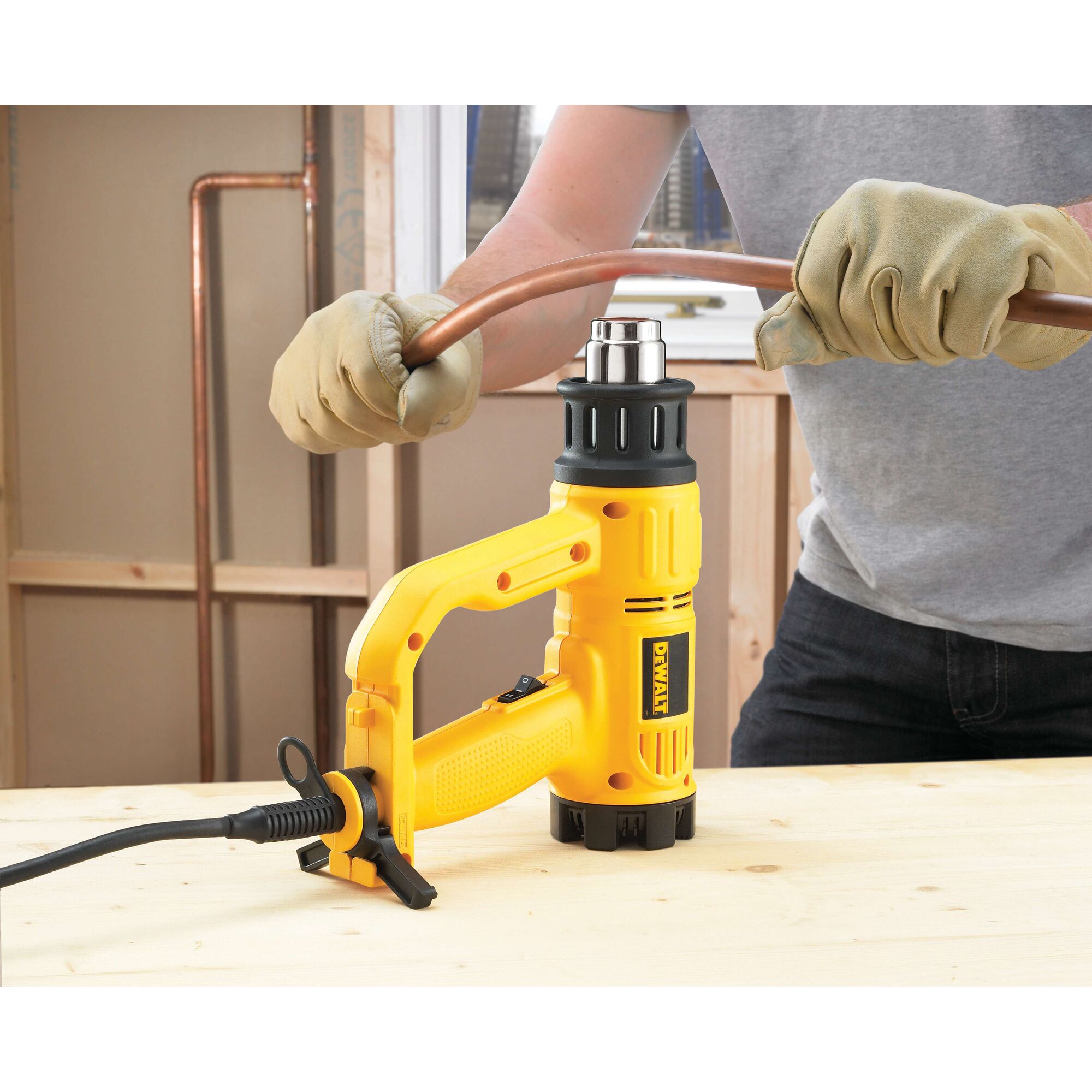 A person wearing work gloves is using a yellow DEWALT heat gun (model D26411) to bend a copper pipe on a wooden workbench in a construction setting.