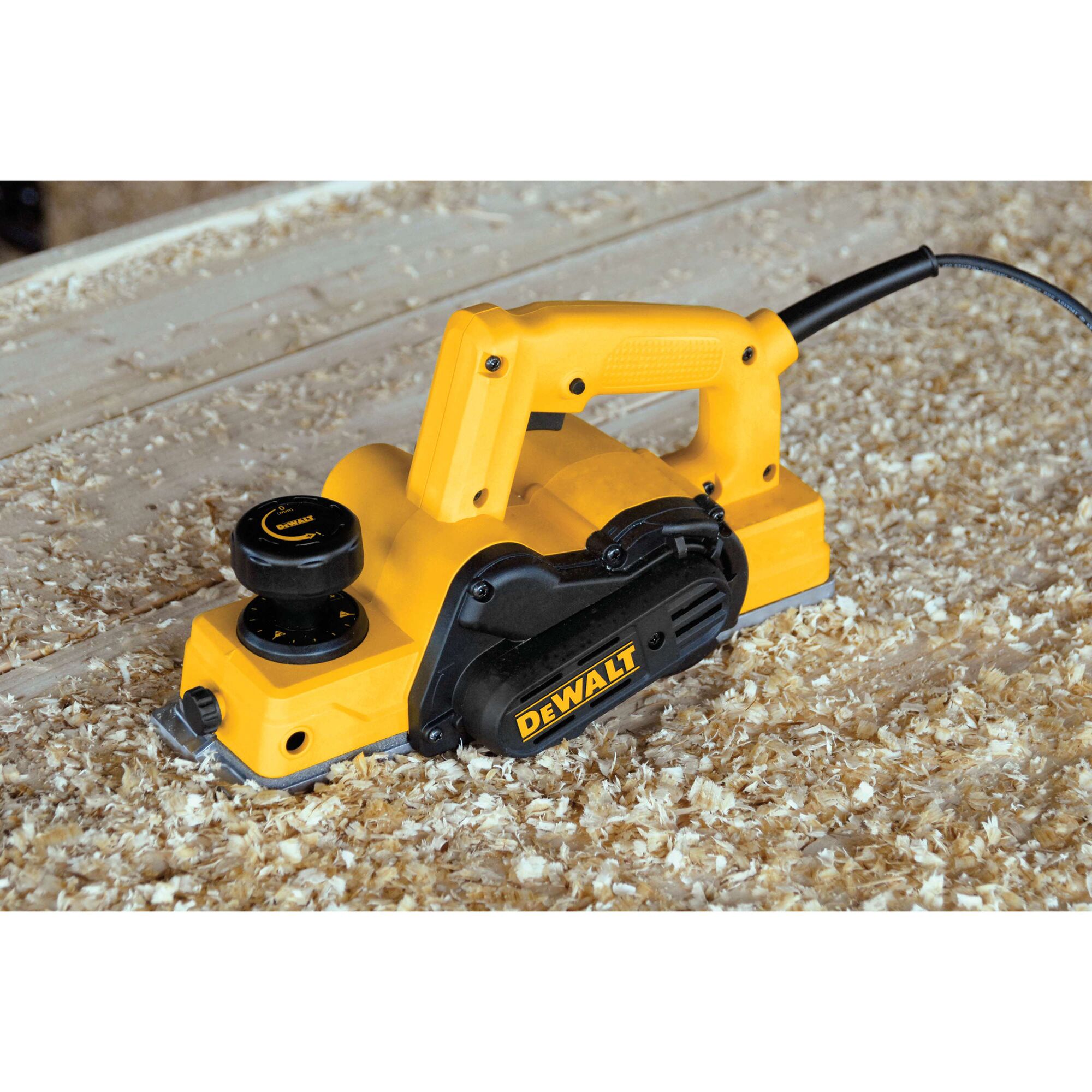 A DEWALT electric hand planer placed on a wooden surface covered with wood shavings. The planer is yellow and black, and is plugged in with a visible power cord.
