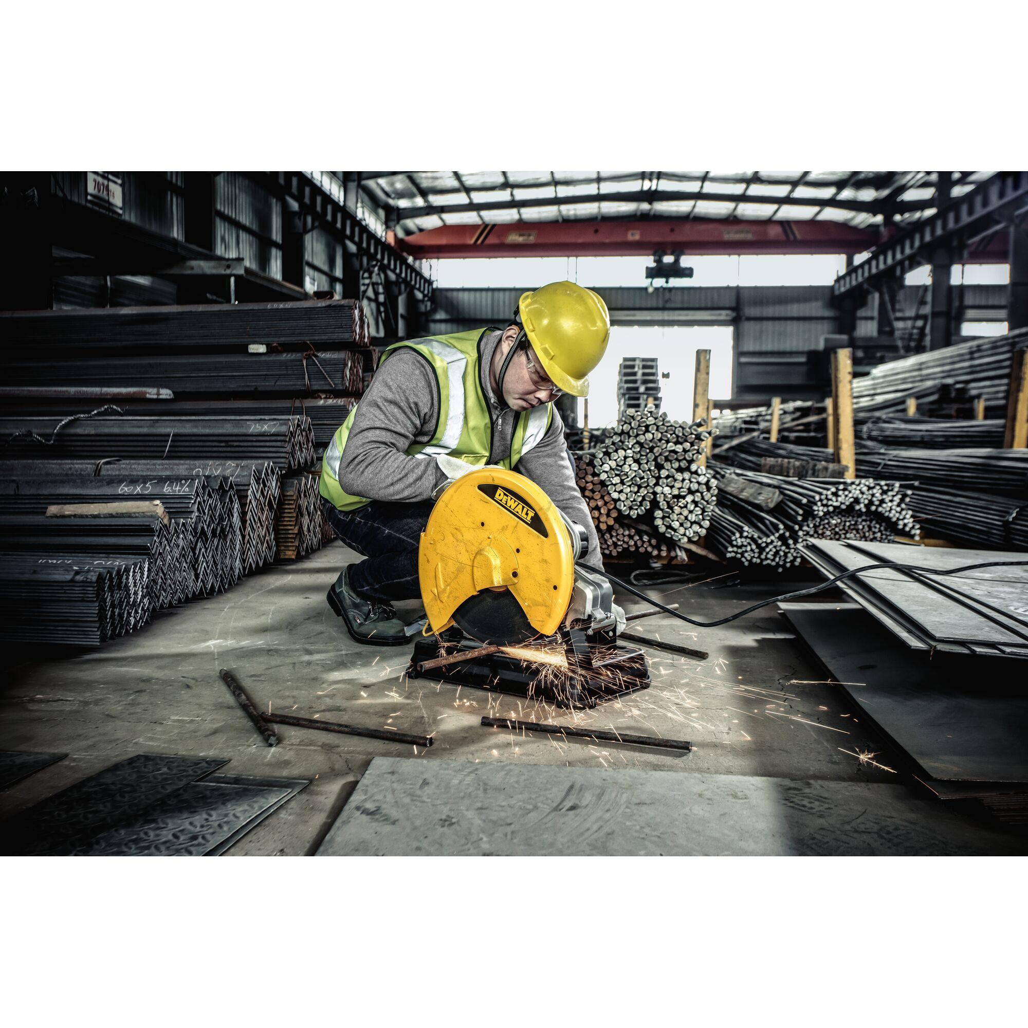 A worker in a yellow hard hat and reflective vest is using a yellow DeWALT metal cutting saw to cut metal rods in an industrial warehouse filled with stacks of steel materials. Sparks are flying from the cutting area.