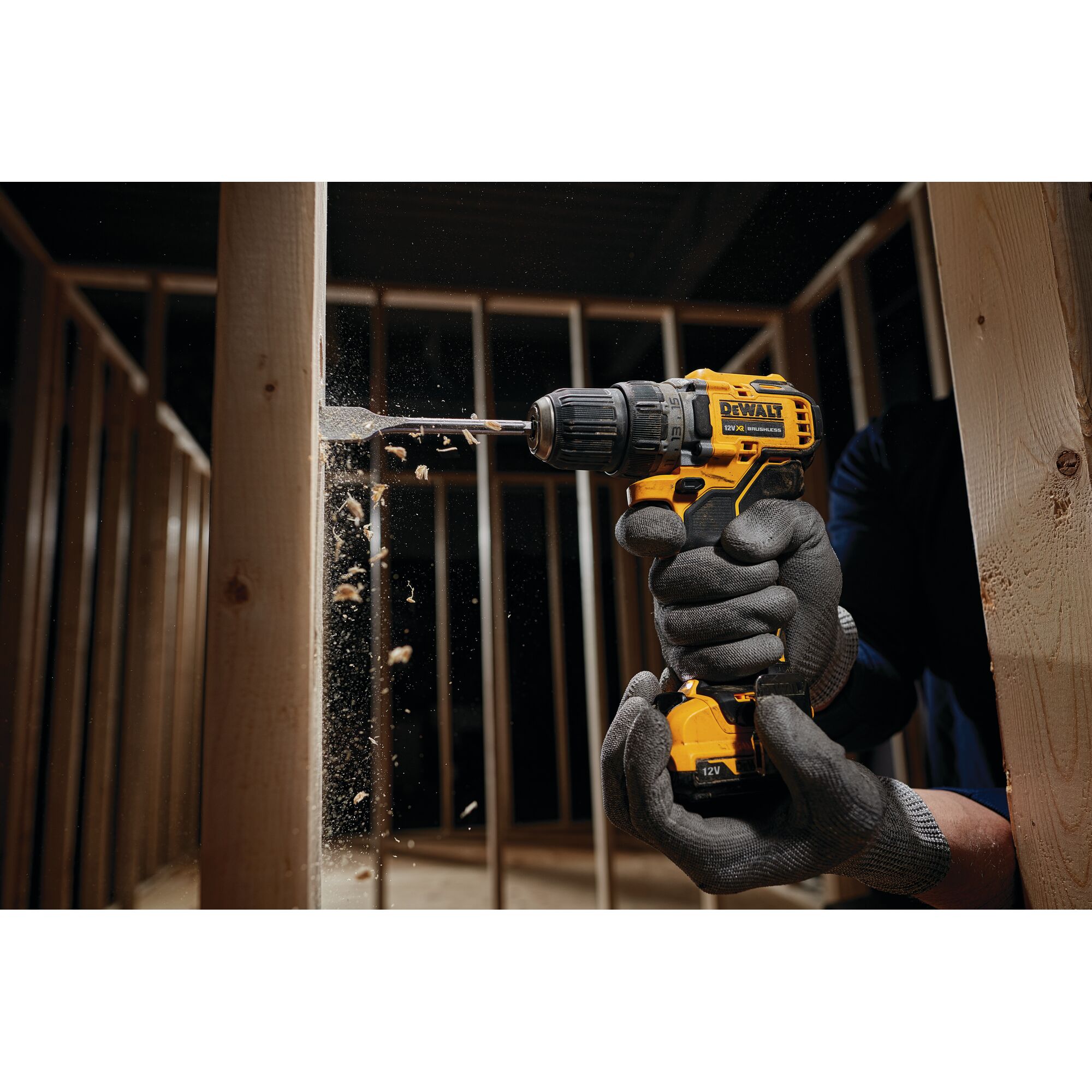 A person wearing protective gloves uses a yellow DEWALT cordless drill to drill into a wooden beam inside a framed structure. Wood chips are visible flying from the drilling process.