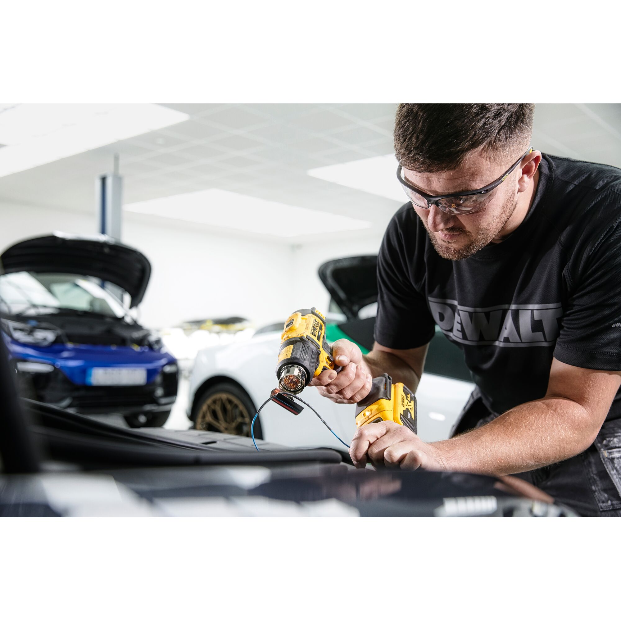 A person using a DEWALT cordless heat gun (model DCE530N) on a car part, with the face blurred for privacy. The individual is wearing a DEWALT-branded shirt and is working in a garage with cars in the background.