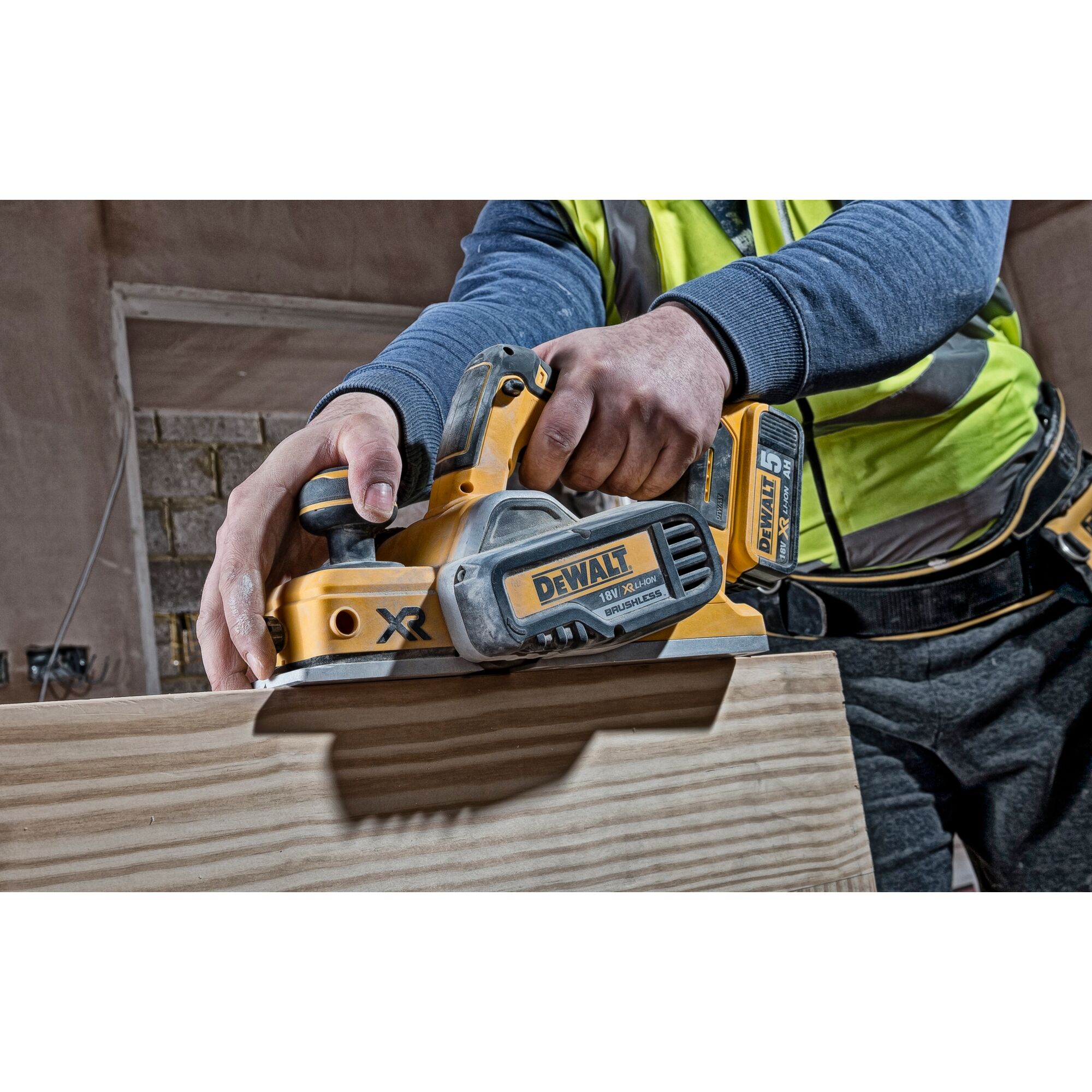 A person using a DEWALT DCP580N cordless planer to smooth the surface of a wooden board. The tool is yellow and black, with visible branding and battery.