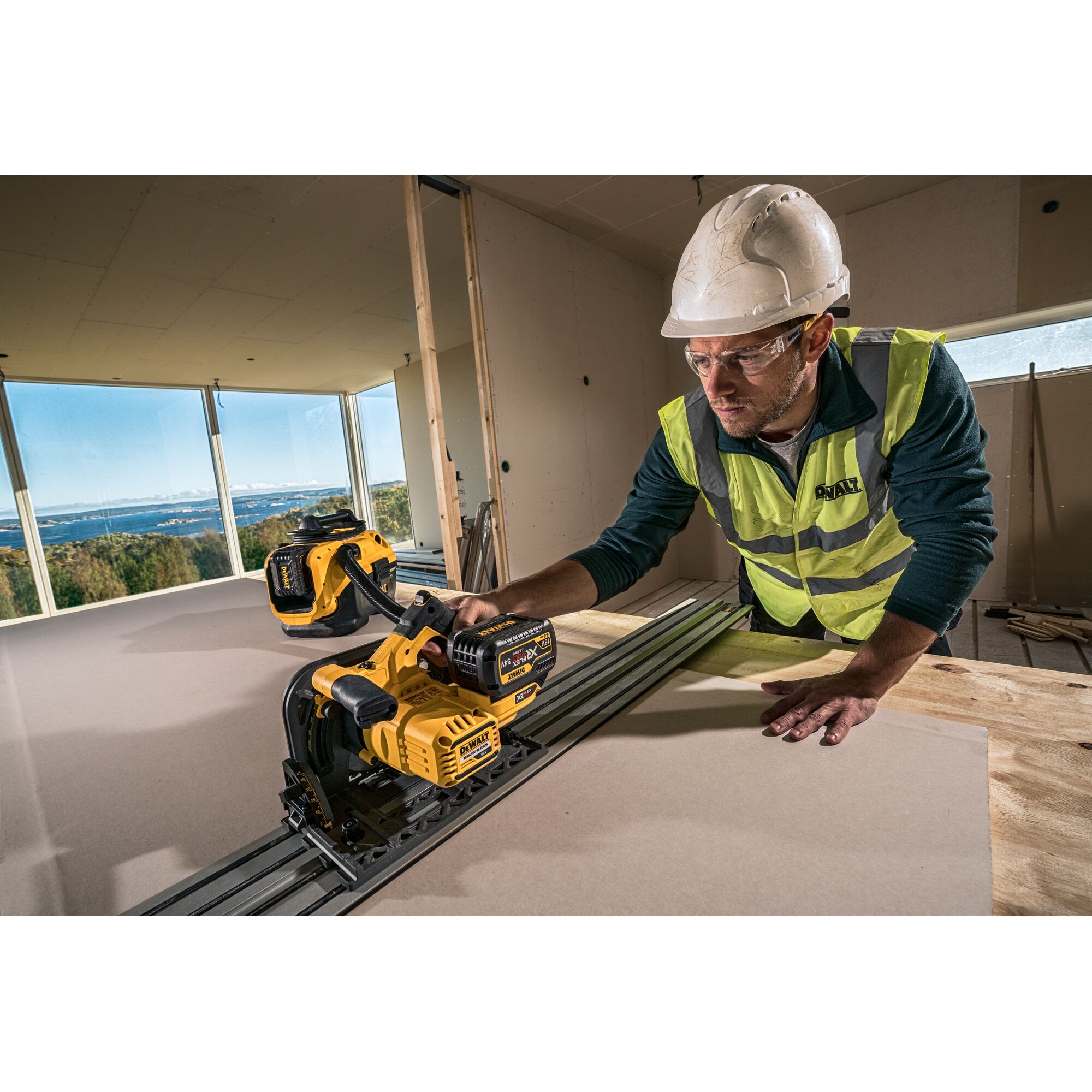 Worker using DEWALT cordless track saw DCS520NT on wood board in modern construction site with safety gear.
