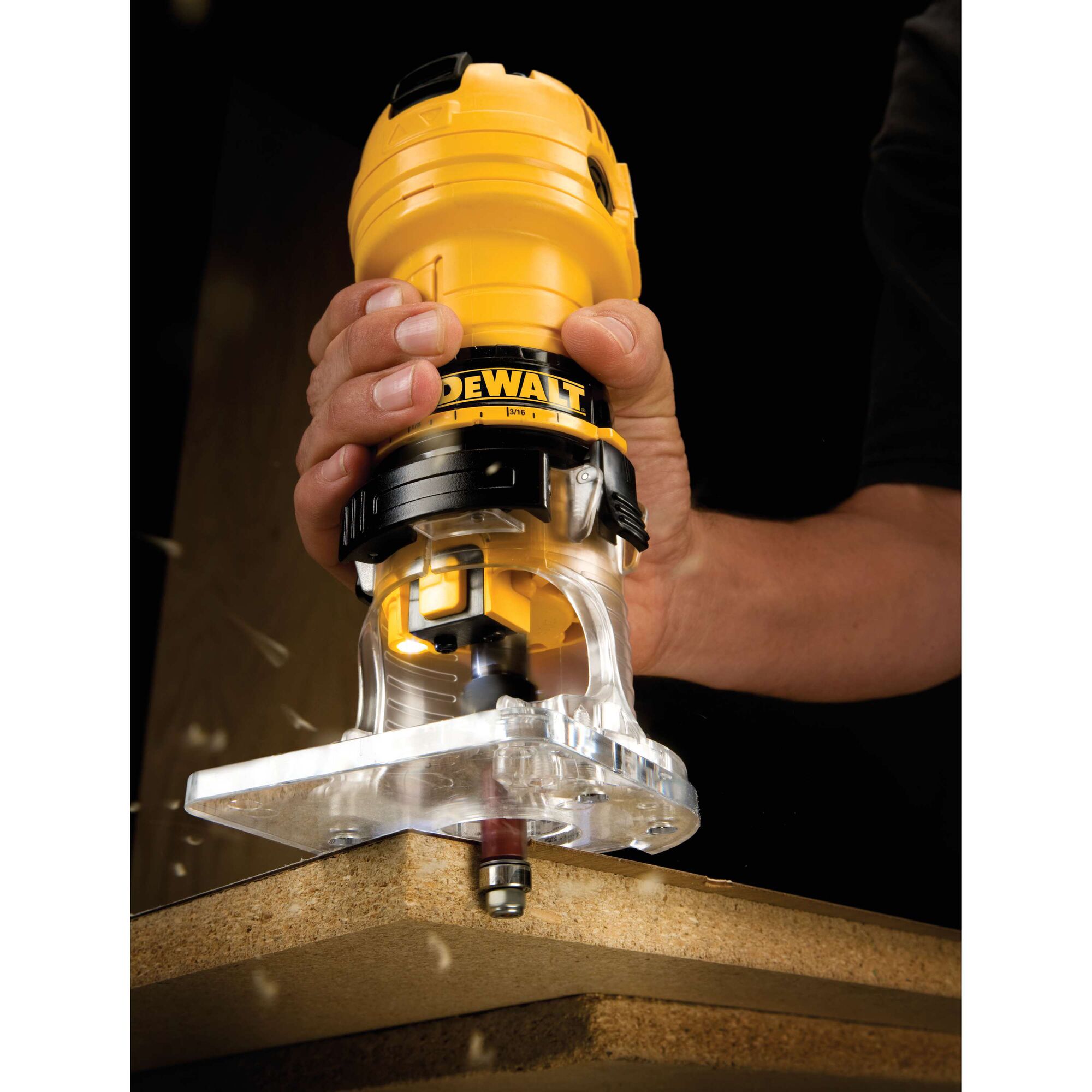 A close-up view of a person using a yellow DEWALT hand router to trim the edge of a wooden board. The router is held firmly and there are wood particles visible as it cuts.