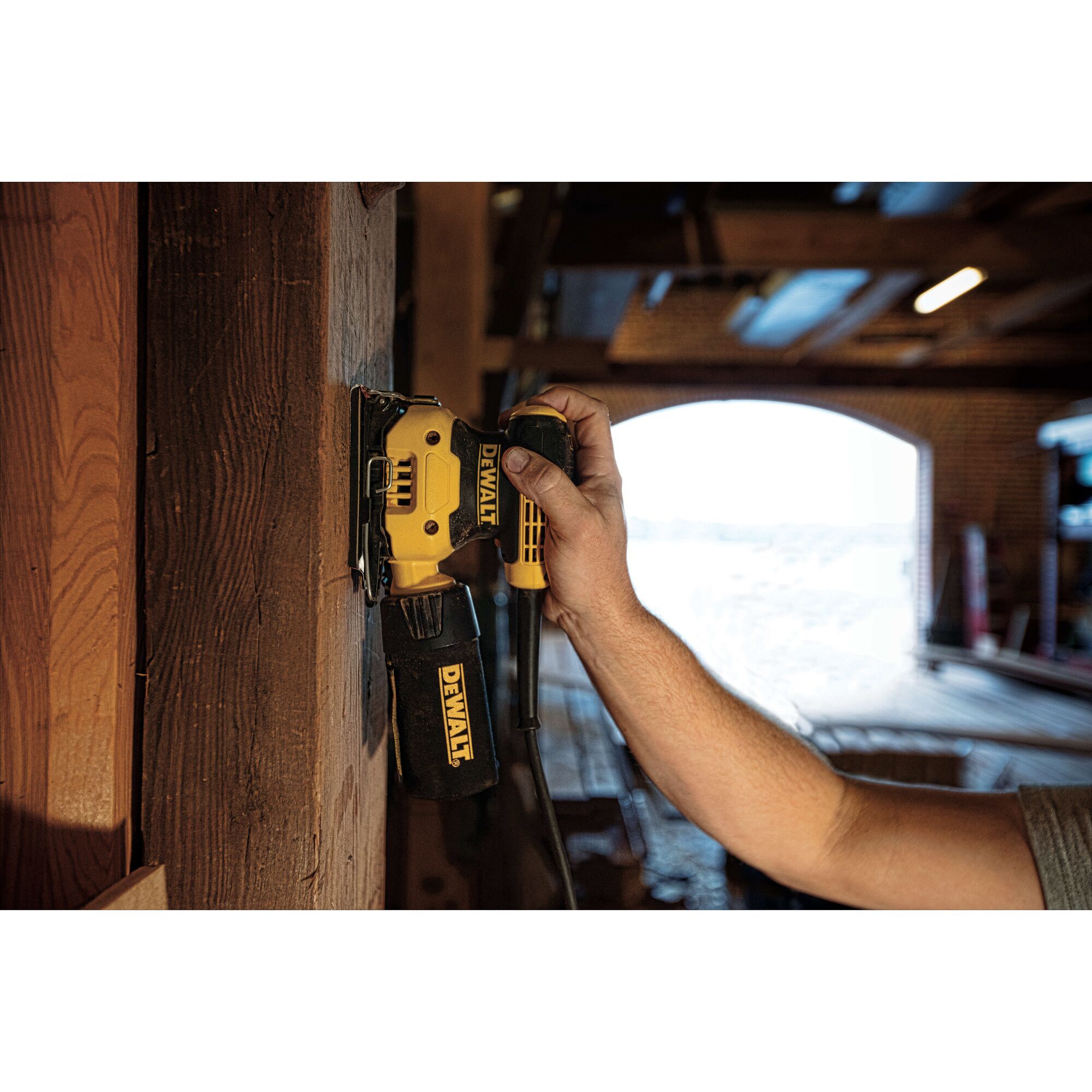 A person using a DEWALT electric sander on a wooden surface inside a workshop or garage. The sander is yellow and black, and the person's hand is holding it against the wood.