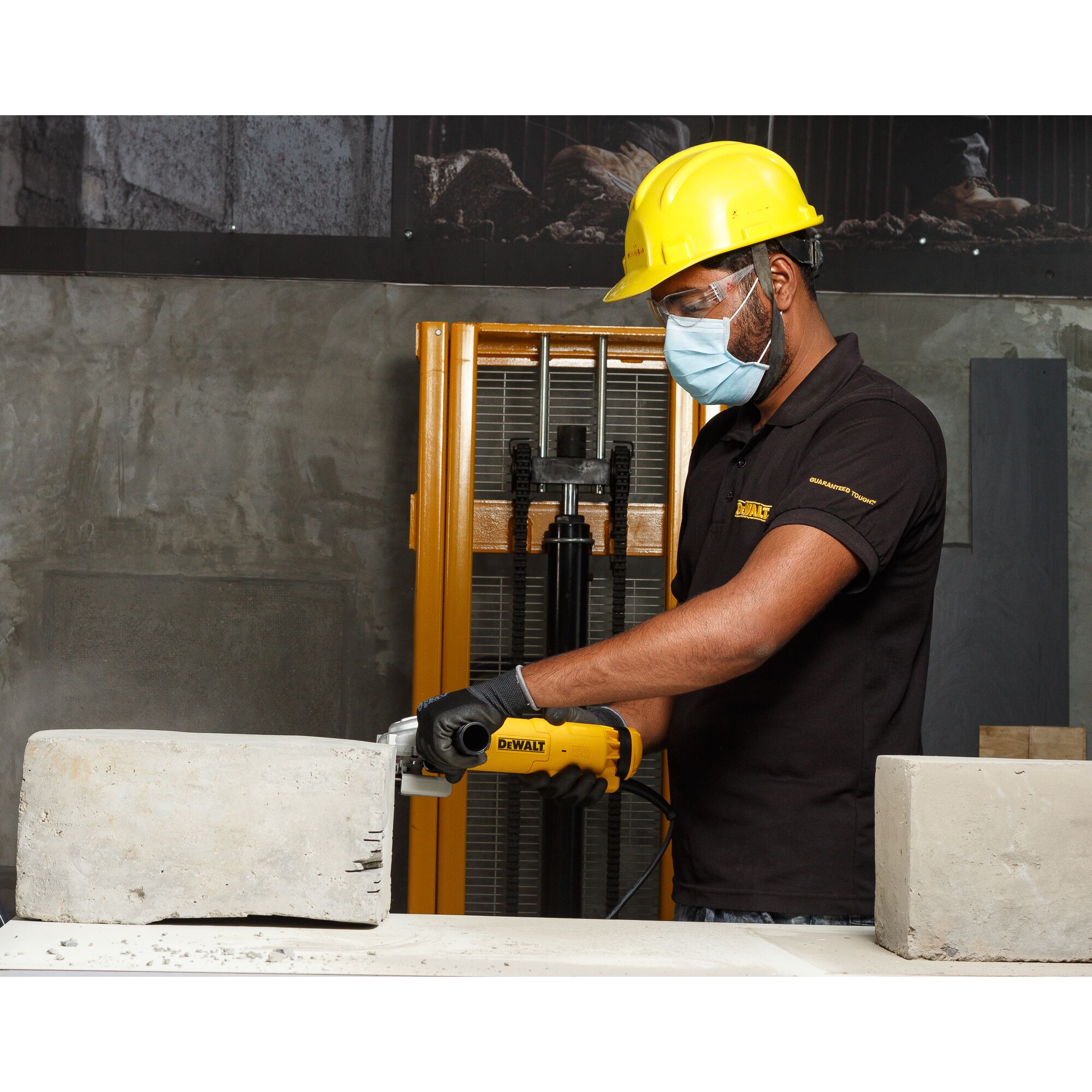A person wearing a yellow safety helmet and gloves is using a DEWALT DWE8300S power tool to cut or grind a concrete block in an industrial environment. The person's face is blurred for privacy.