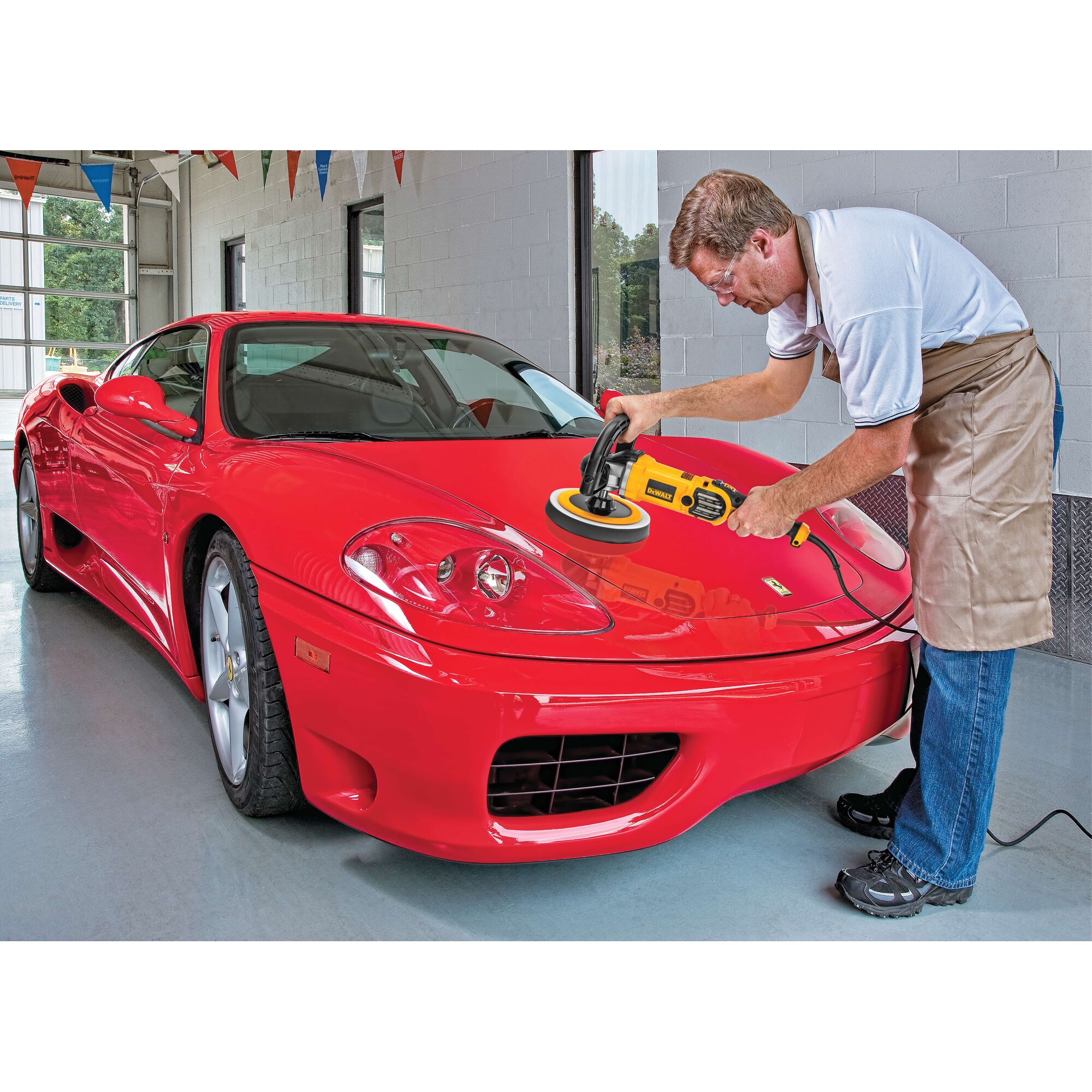 A person using a DEWALT DWP849X polisher to buff the hood of a shiny red sports car inside a garage. The person is wearing an apron and is focused on the polishing process.