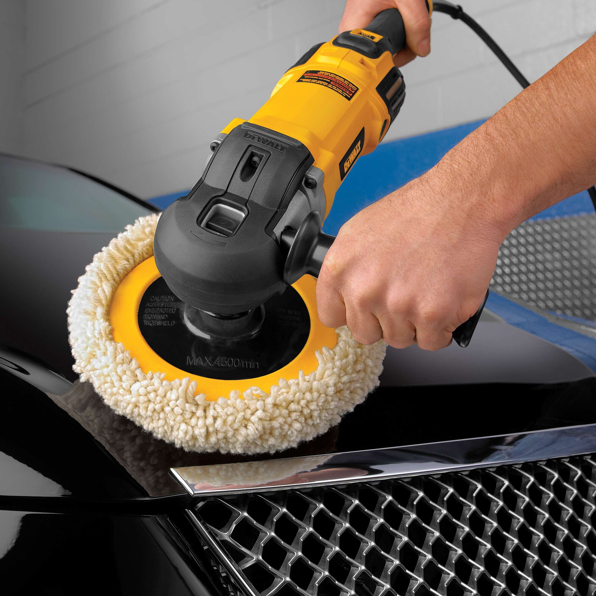A person uses a yellow electric car polisher with a fluffy buffing pad to polish the surface of a black car hood near the grille.