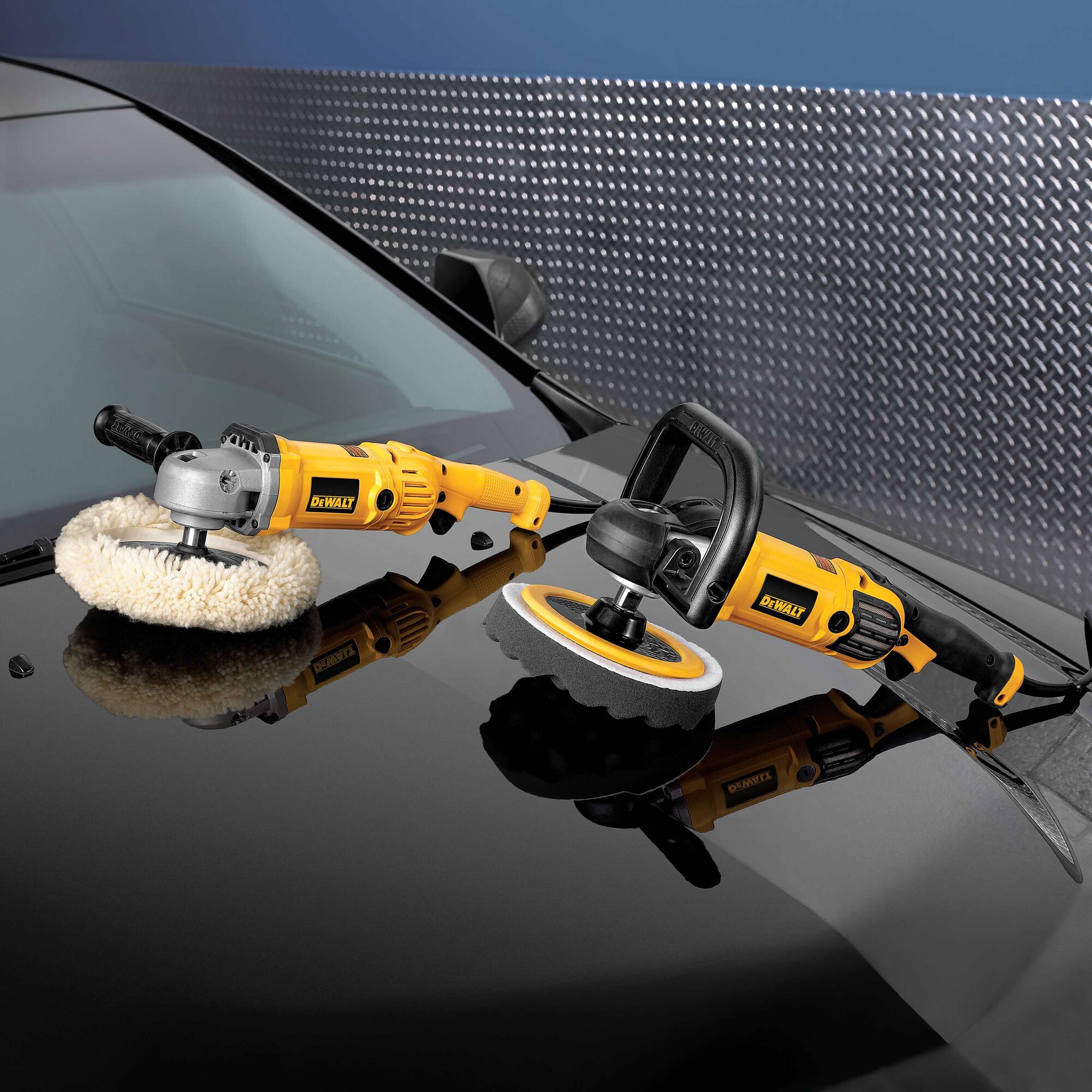 Two DEWALT electric polishers are placed on the hood of a black car. Both tools have yellow and black bodies and are equipped with polishing pads, ready for use. The background includes a metal panel wall.
